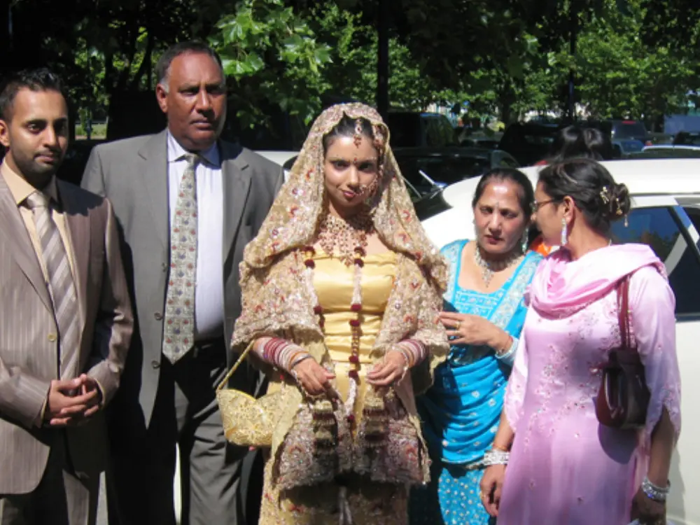 Asian bride in traditional gold embroidered attire standing with family members by wedding car