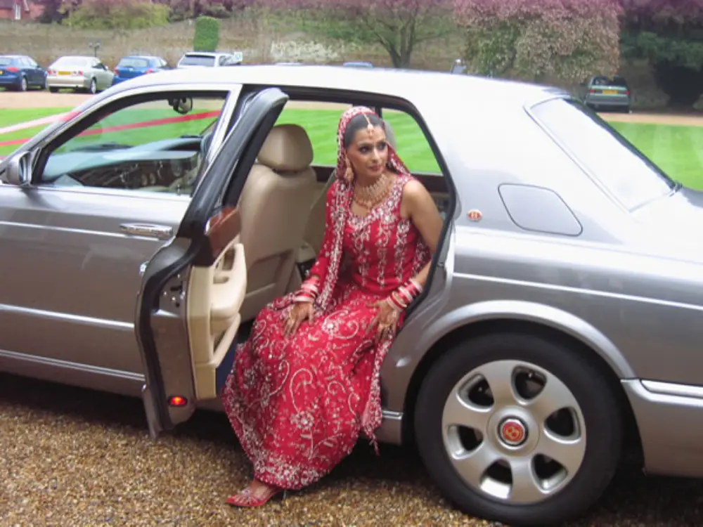 Asian bride in ornate red and gold embroidered dress stepping out of silver Bentley wedding car