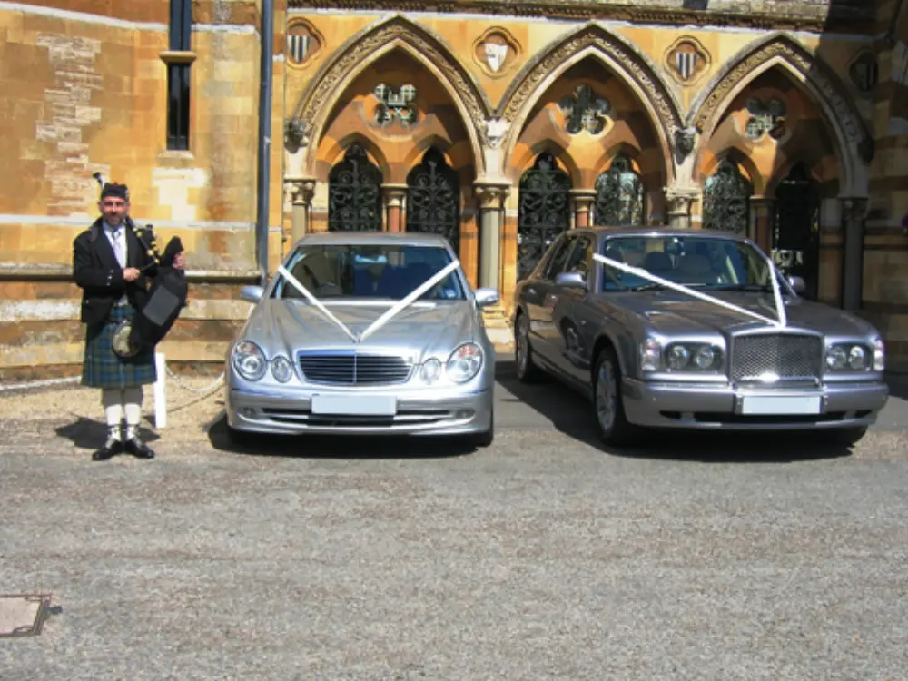 Scottish bagpiper in traditional kilt standing between silver Mercedes and Bentley wedding cars