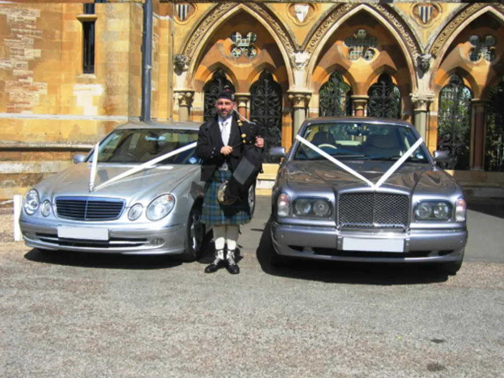 Scottish bagpiper posing between silver Mercedes and Bentley wedding cars decorated with ribbons