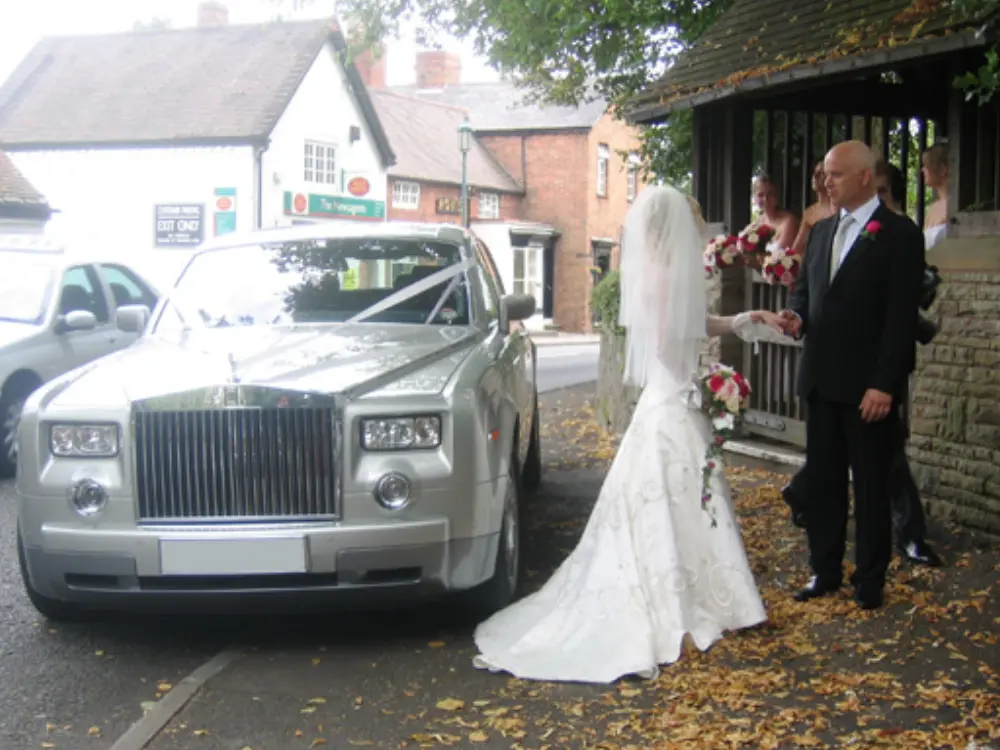 Bride in veil and white gown holding hands with father next to silver Rolls-Royce Phantom wedding car
