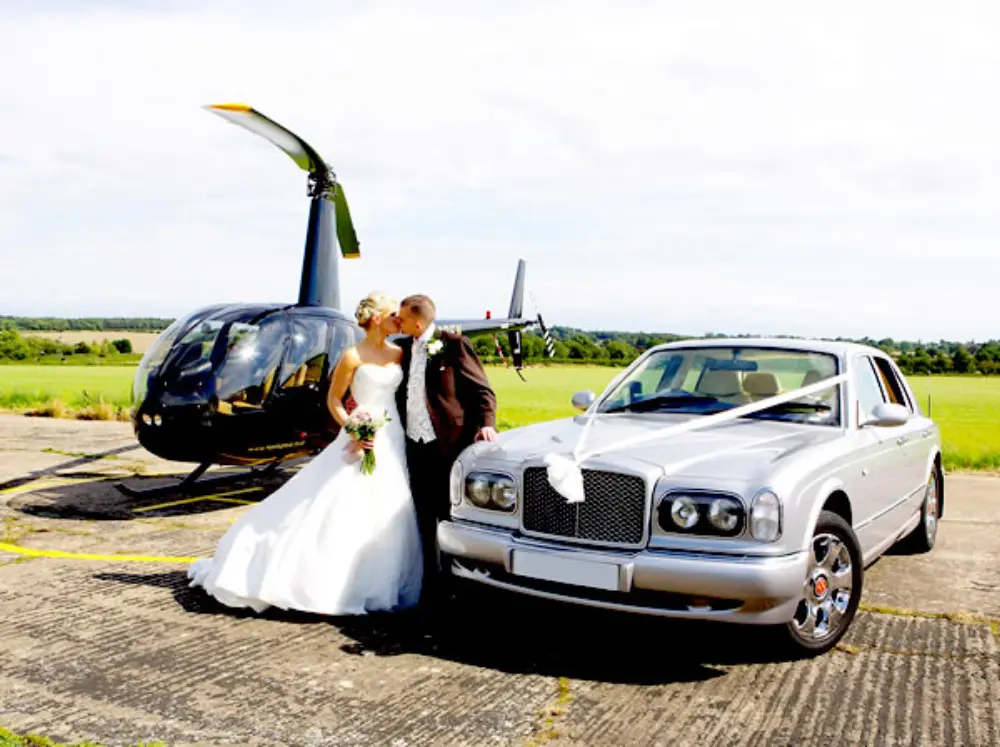 Bride and groom kissing between silver Bentley wedding car and black helicopter on airfield