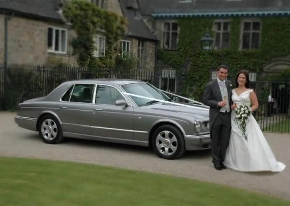 Bride and groom toasting with champagne next to silver Bentley outside historic ivy-covered manor