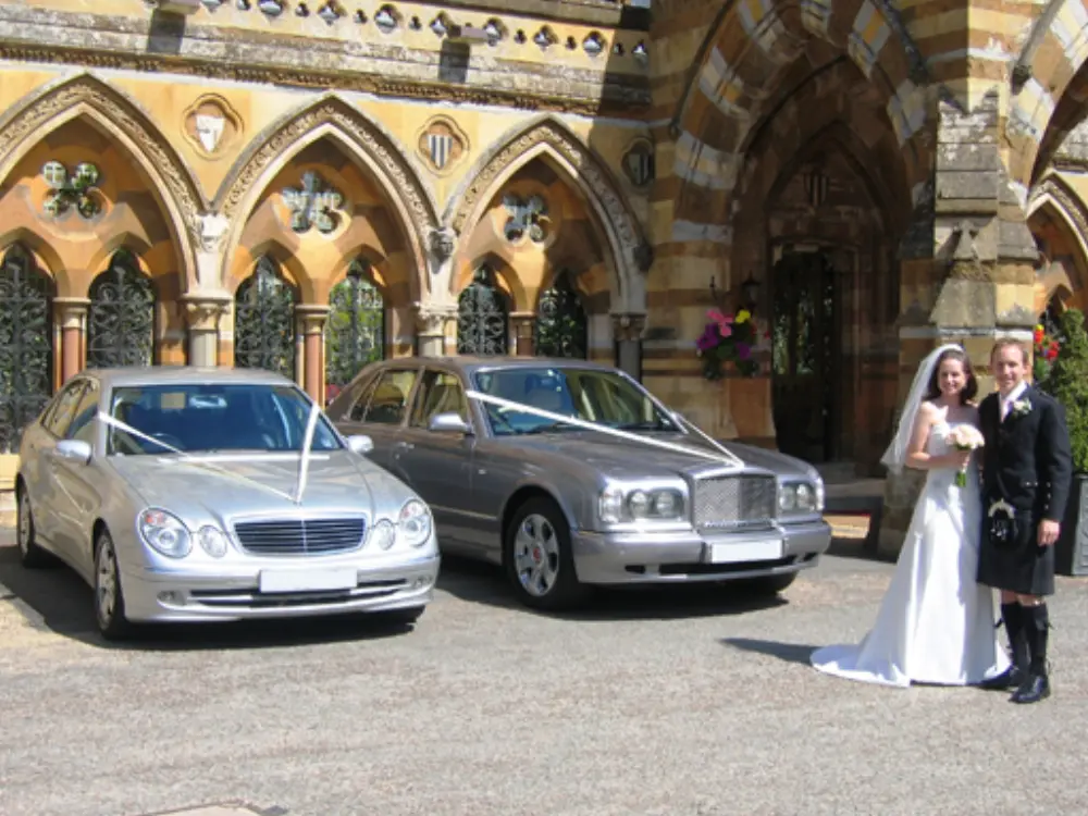 Bride and Scottish groom in kilt posing with silver Mercedes and Bentley wedding cars outside church