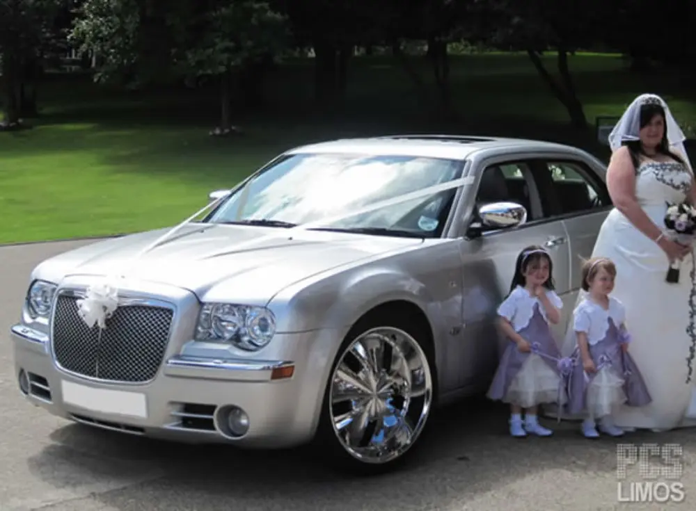 Bride and two flower girls in lilac dresses posing next to silver Chrysler 300C wedding car