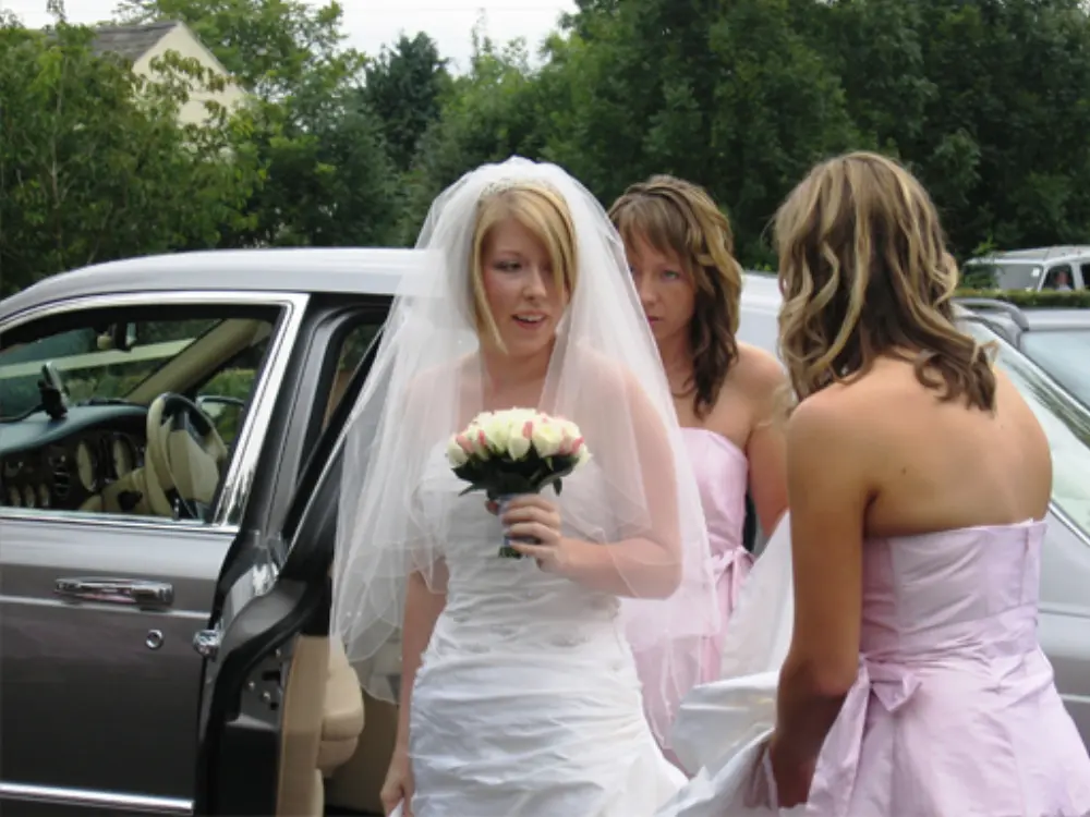 Bride with veil and rose bouquet assisted by bridesmaids in pink dresses stepping out of silver wedding car