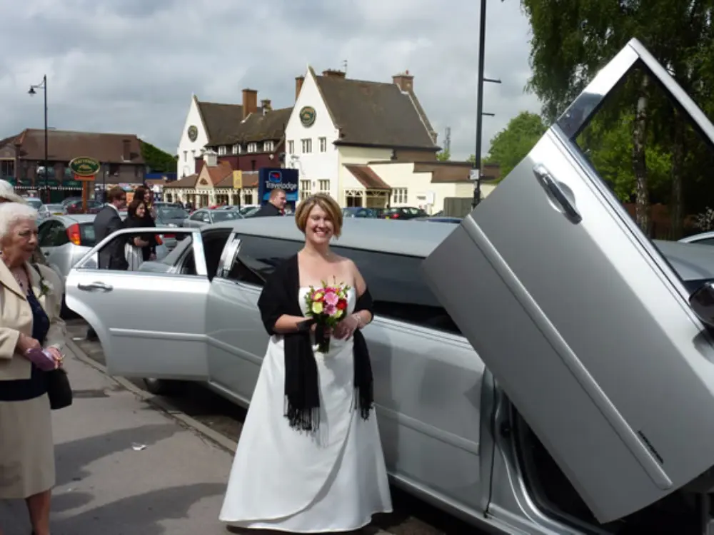 Bride standing next to silver Chrysler stretch limousine with open scissor door
