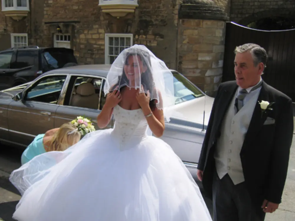 Bride in full ballgown wedding dress with veil and father in morning suit next to silver wedding car