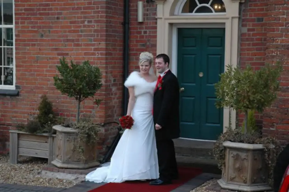 Bride in white fur stole and groom in black tuxedo standing on red carpet outside venue
