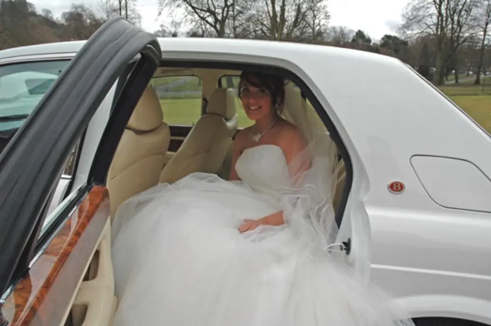 Bride in large white wedding gown seated in back of white Bentley wedding car