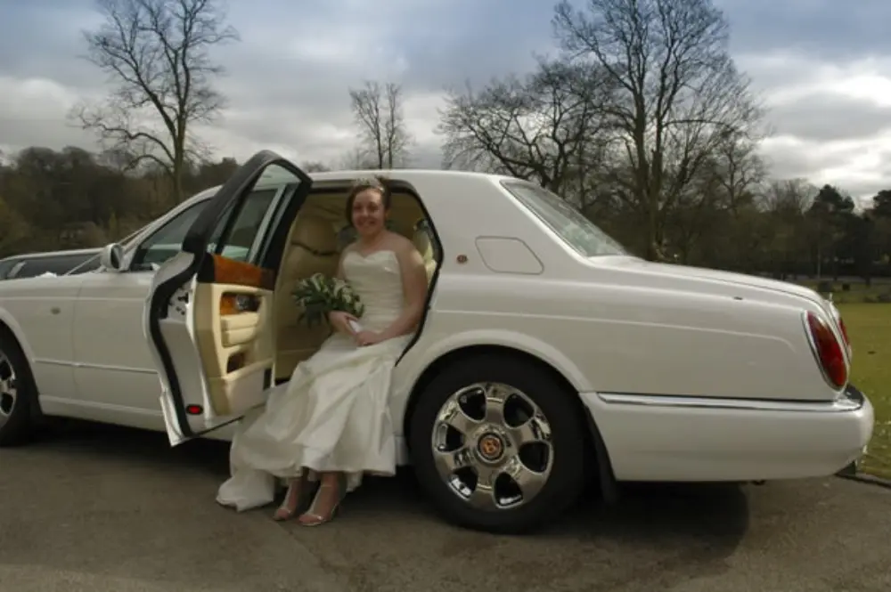 Bride in strapless gown seated at open rear door of white Bentley luxury car