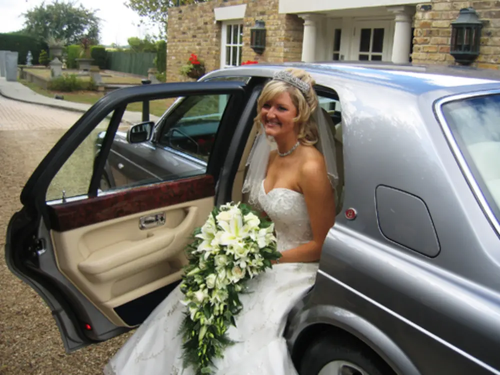 Bride wearing tiara with cascading floral bouquet seated in rear of silver Bentley car