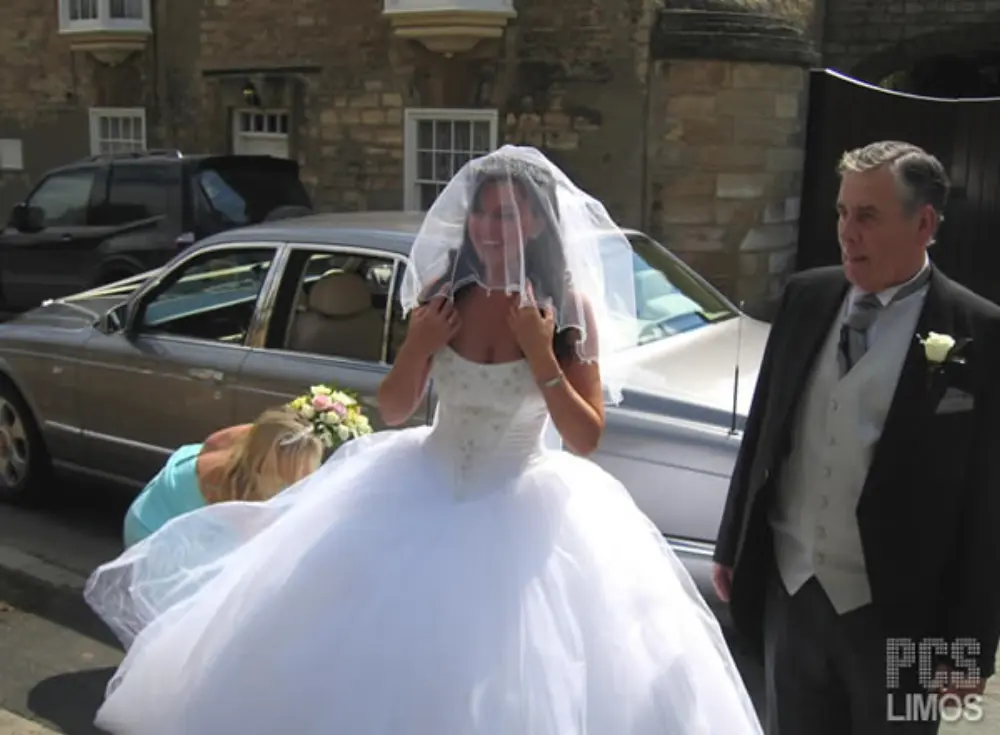 Bride with veil and voluminous gown standing with father beside silver luxury wedding car