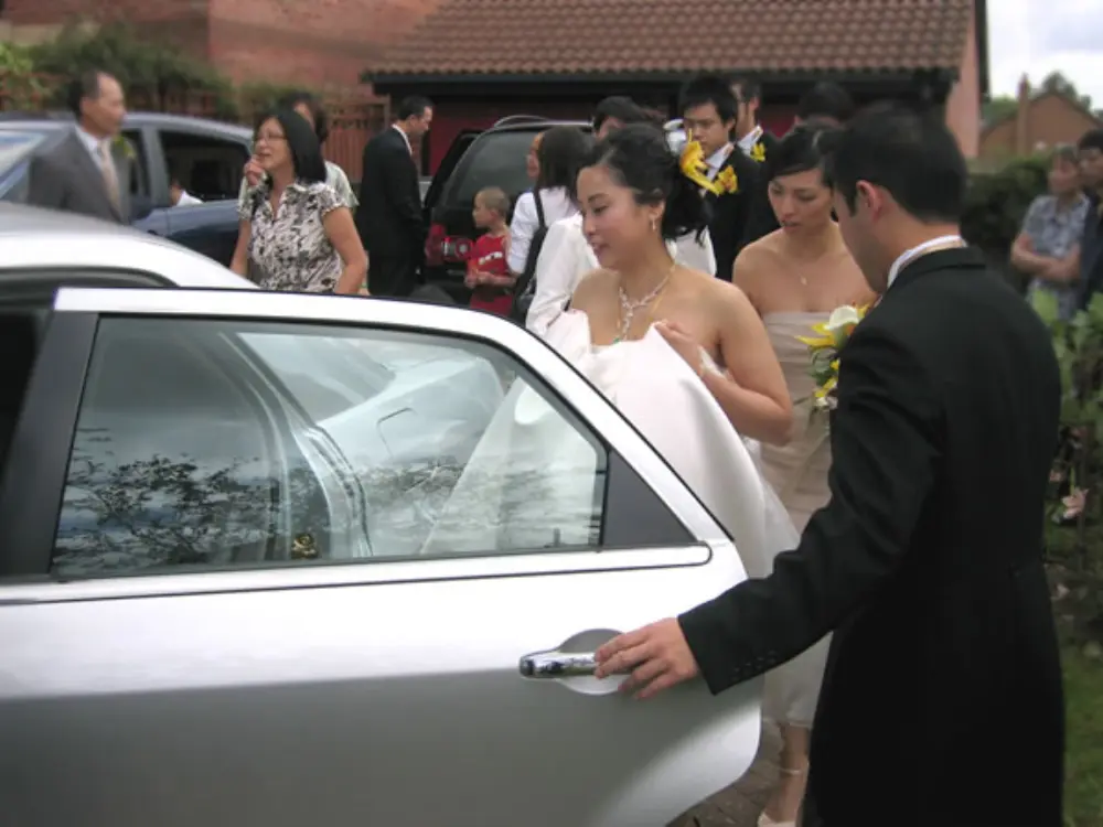 Groom opening silver wedding car door for bride in white dress with guests gathered outside