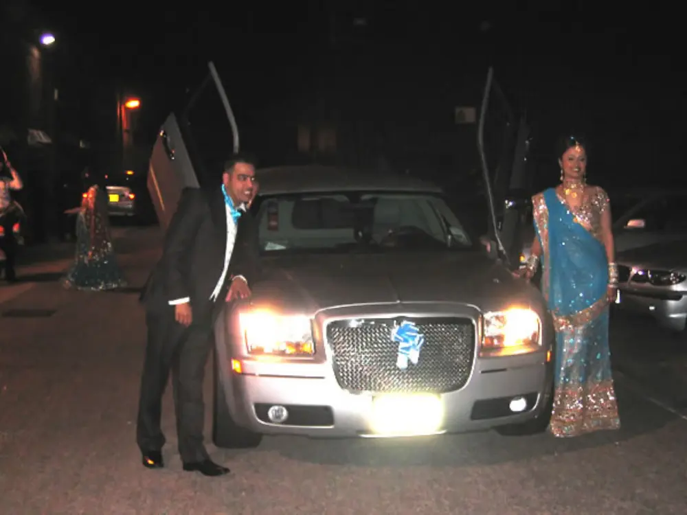 Groom and Indian bride in blue sari posing by illuminated silver Chrysler limousine at night