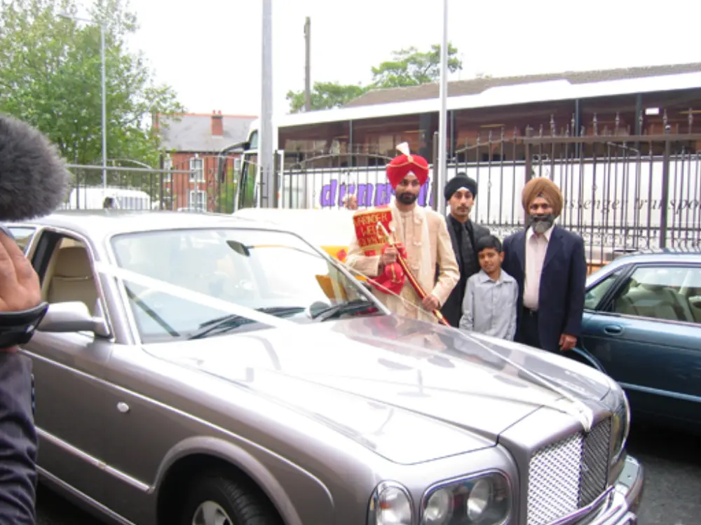 Sikh groom in red turban and family posing in front of ribbon-adorned silver Bentley wedding car