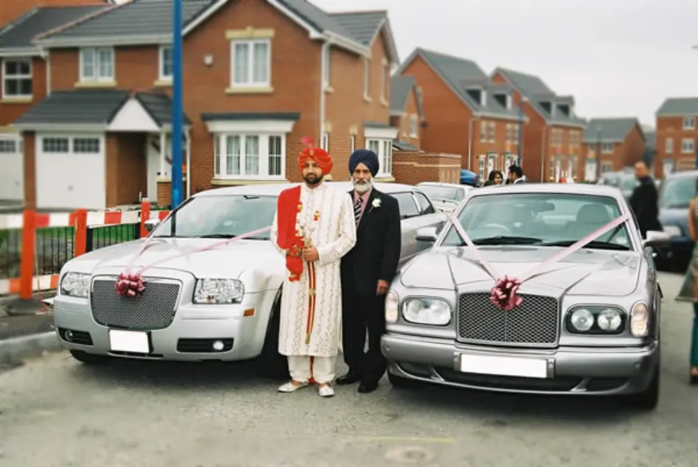 Sikh groom in red turban and father standing between silver Chrysler stretch limo and silver Bentley