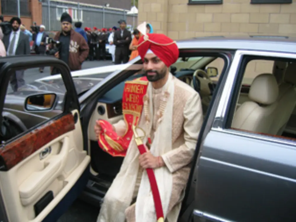Sikh groom in cream sherwani and red turban holding ceremonial sword beside luxury car