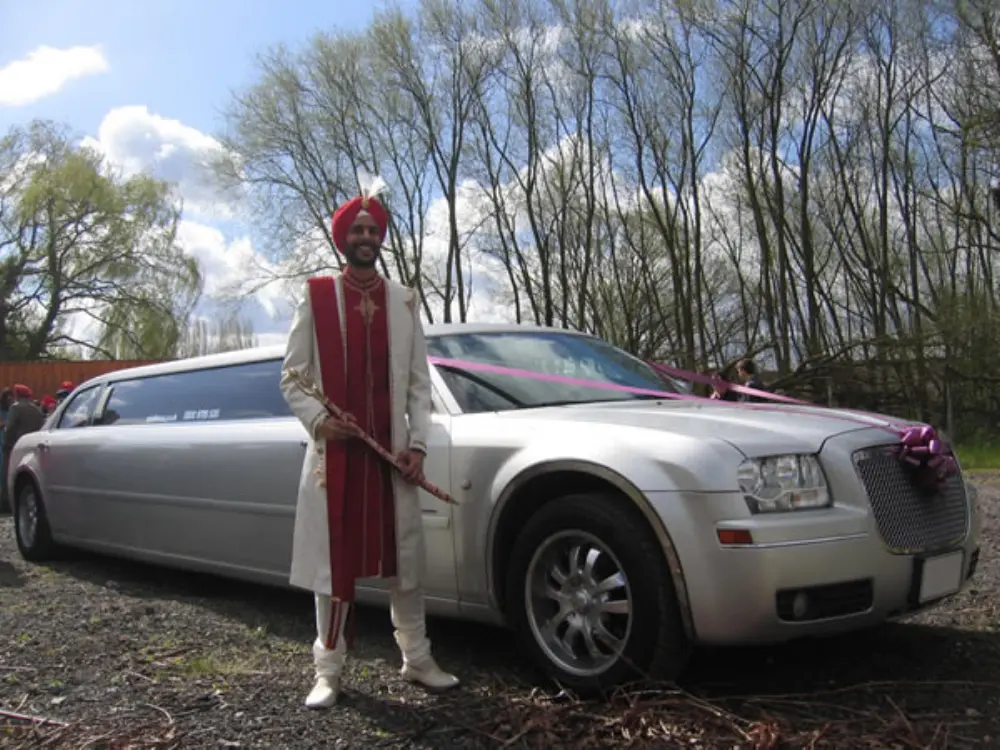 Sikh groom in traditional cream attire with red turban posing beside silver Chrysler stretch limousine