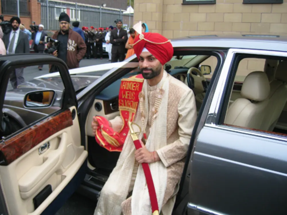 Sikh groom in traditional cream sherwani and red turban holding sword stepping out of silver Bentley