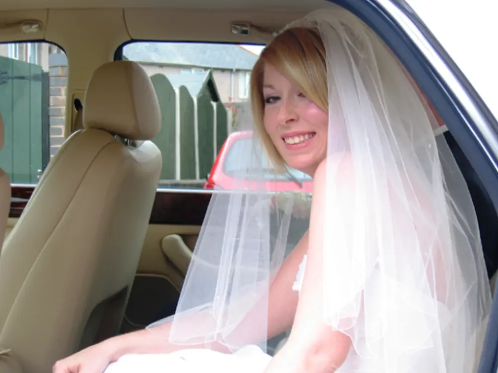 Smiling blonde bride with veil sitting in cream leather back seat of luxury wedding car