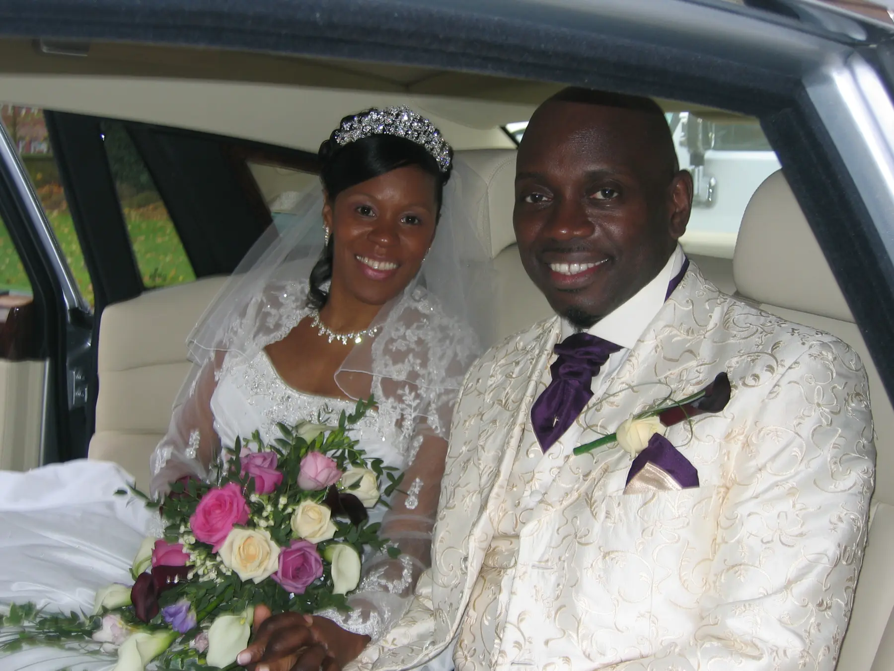 Smiling bride in lace gown and groom in cream suit holding hands inside Rolls-Royce wedding car