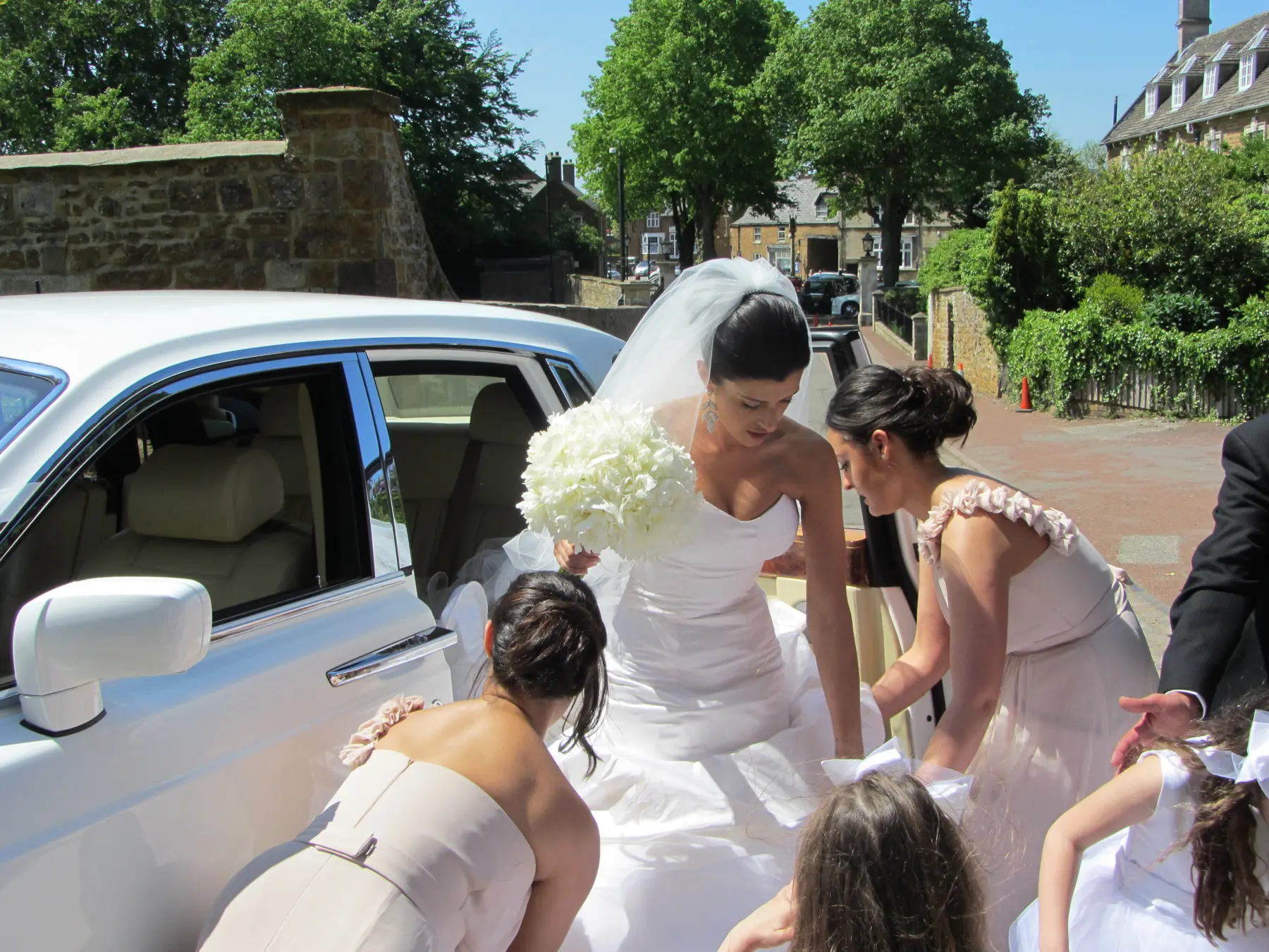 Bride holding white bouquet assisted by bridesmaids stepping out of white Rolls-Royce Phantom
