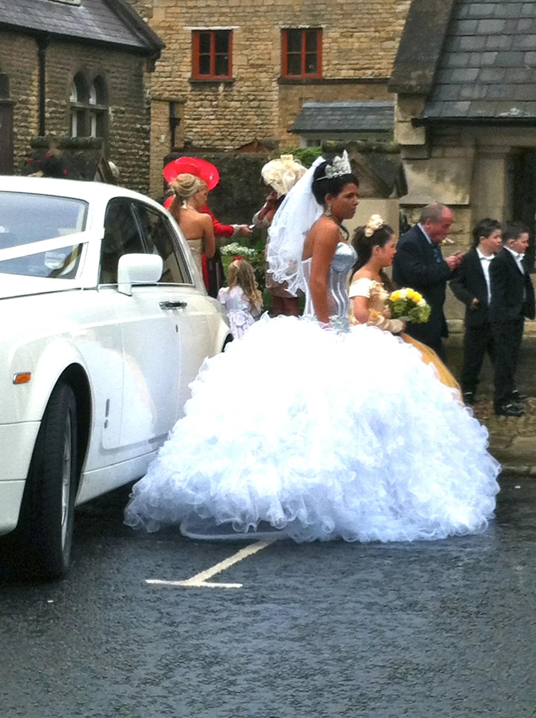 Gypsy bride in enormous ruffled ballgown standing beside white Rolls-Royce Phantom wedding car