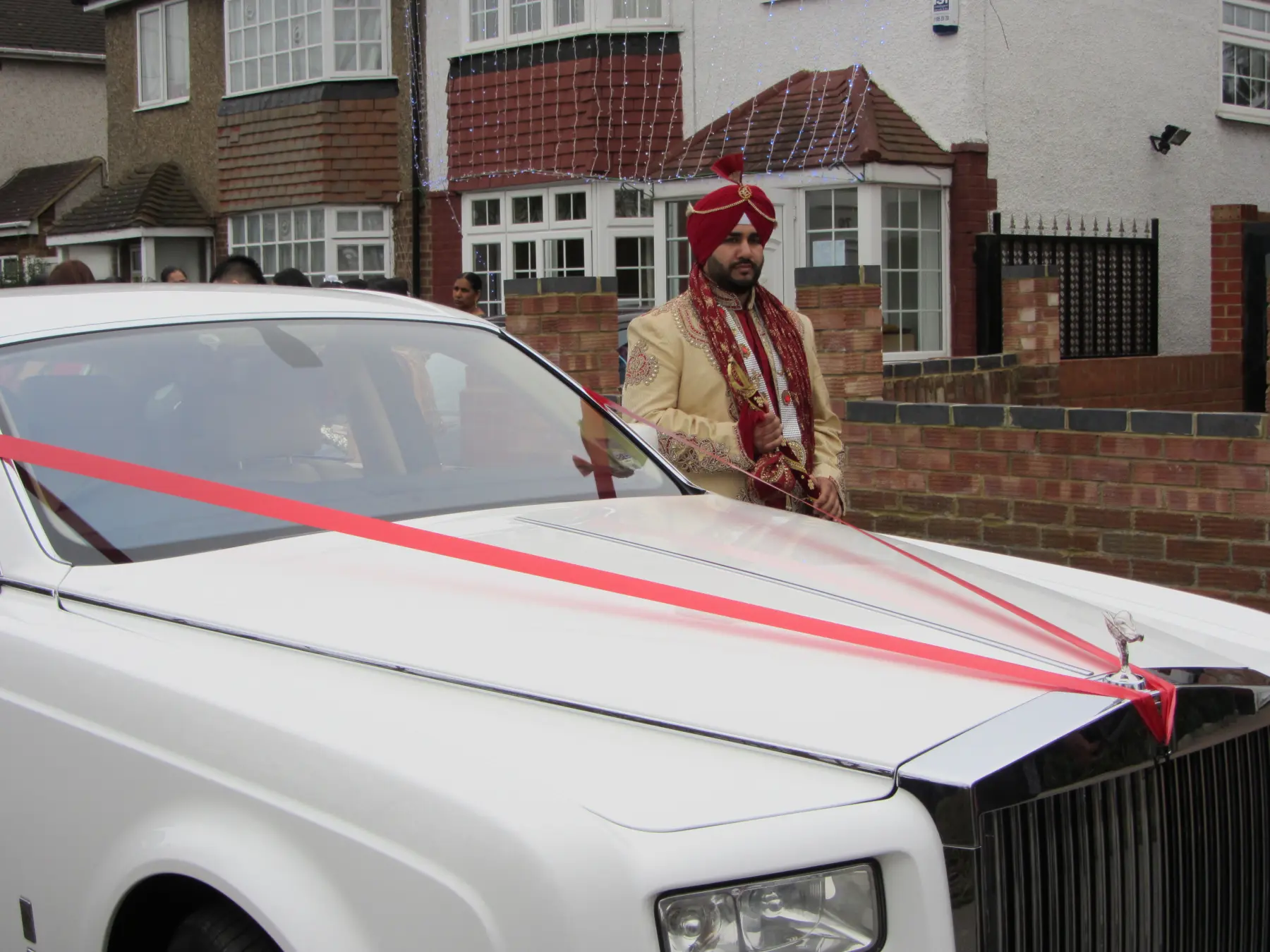Sikh groom in traditional red turban standing beside white Rolls-Royce Phantom decorated with red ribbons