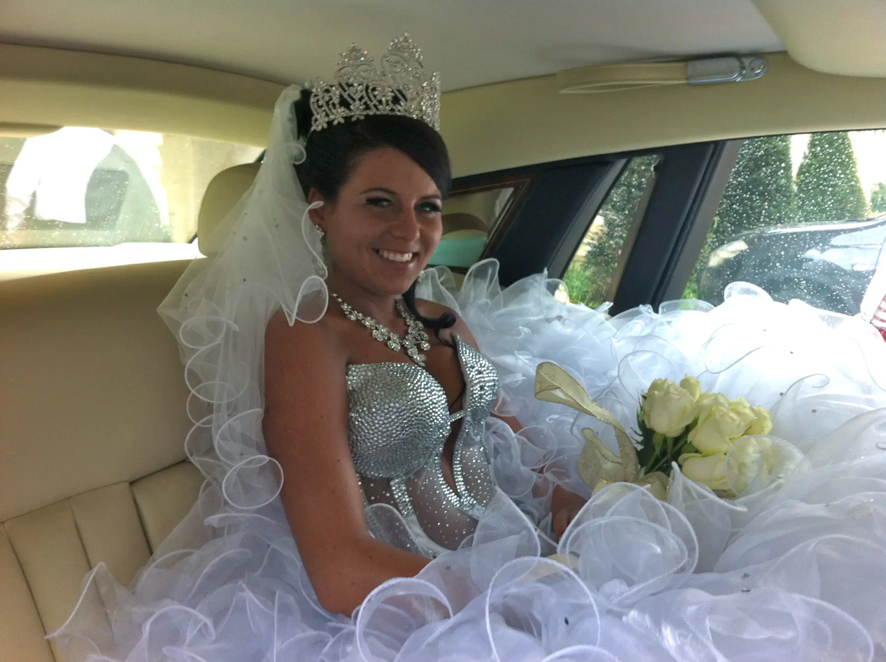 Smiling gypsy bride wearing tall crystal crown and jewel corset seated inside luxury wedding car