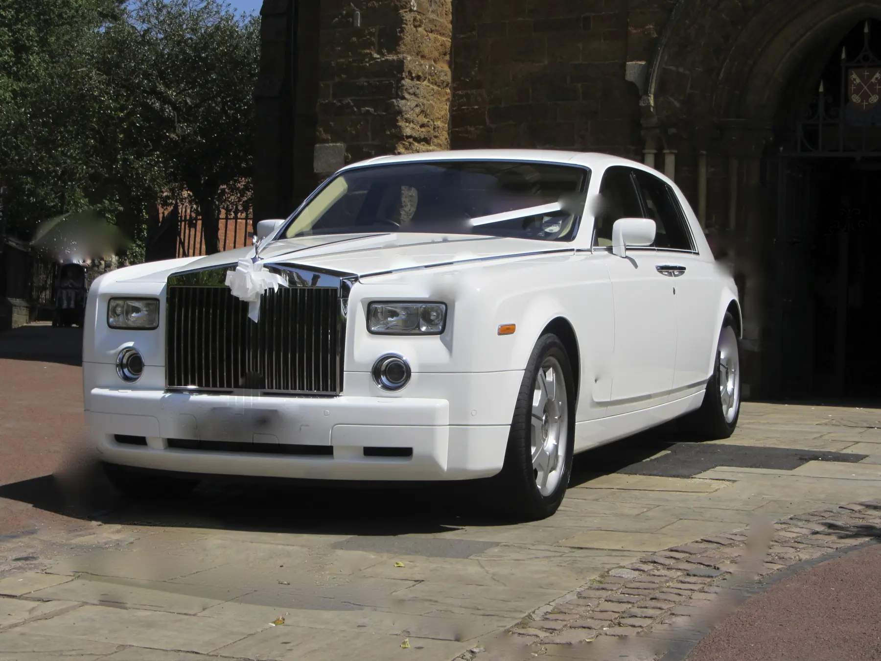 White Rolls-Royce Phantom wedding car parked under sunny stone church entrance archway