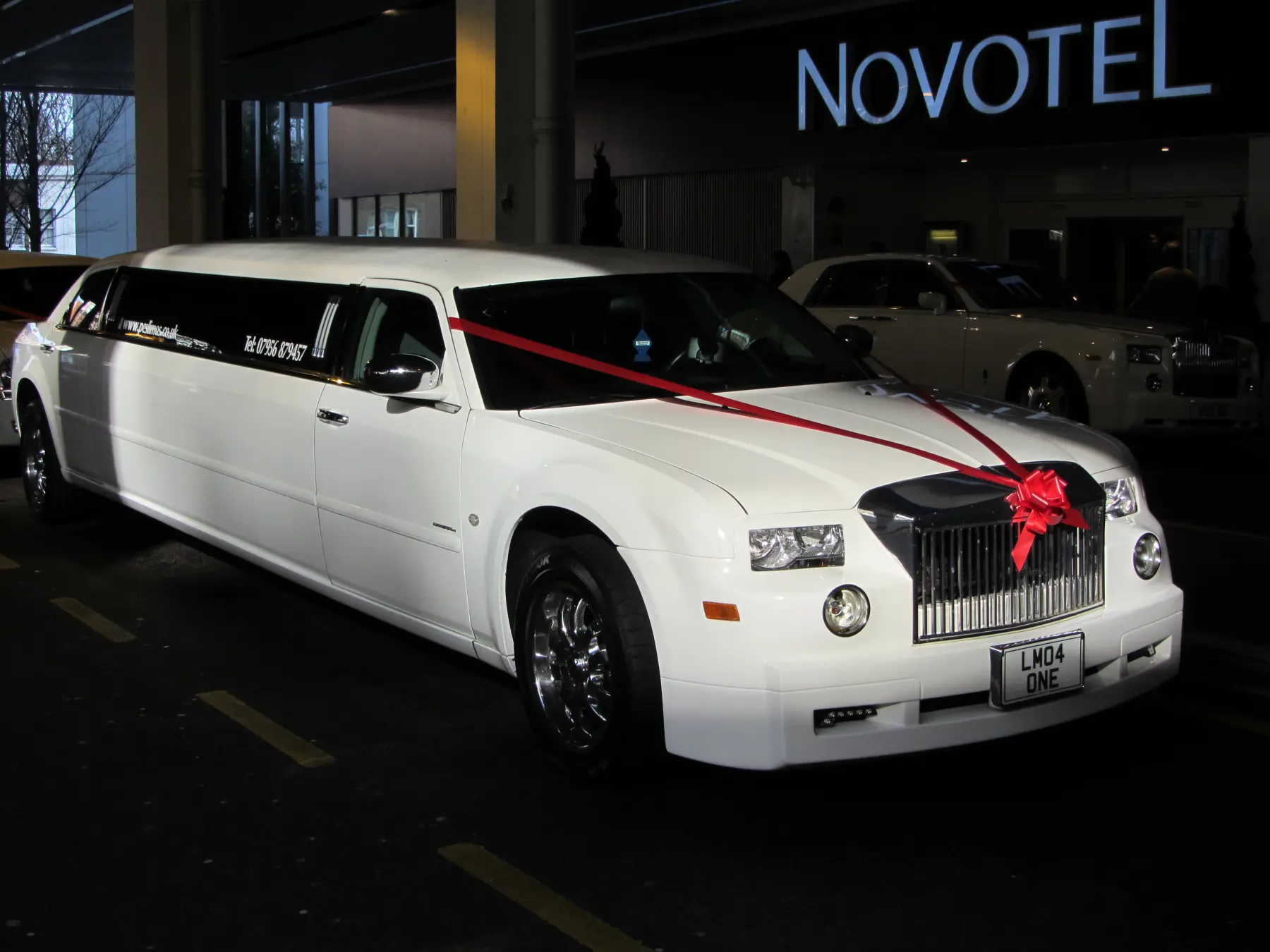 White Rolls-Royce style stretch limousine with red ribbon and bow parked outside Novotel hotel