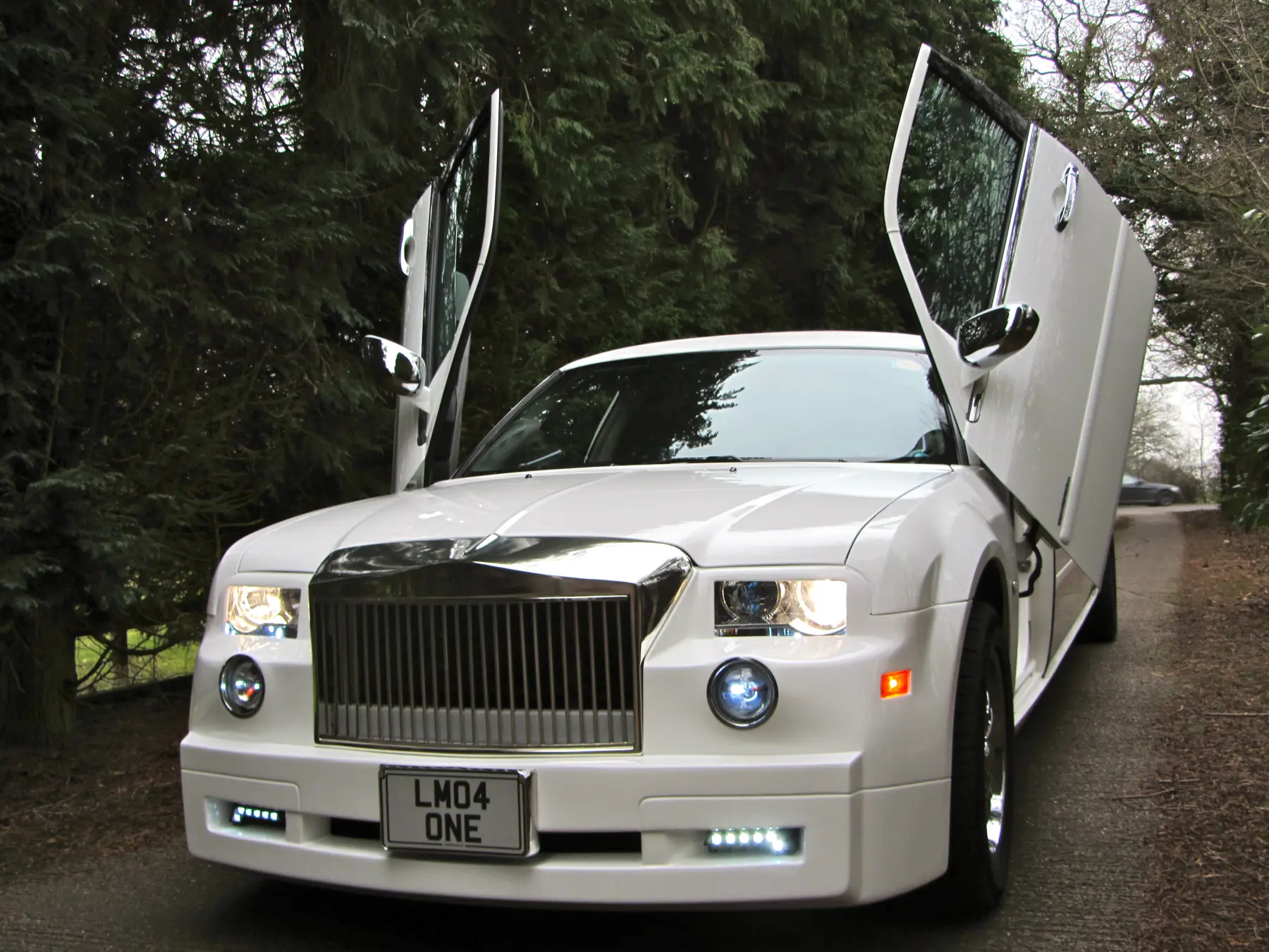 White Rolls-Royce style stretch limousine with scissor doors raised on tree-lined country lane