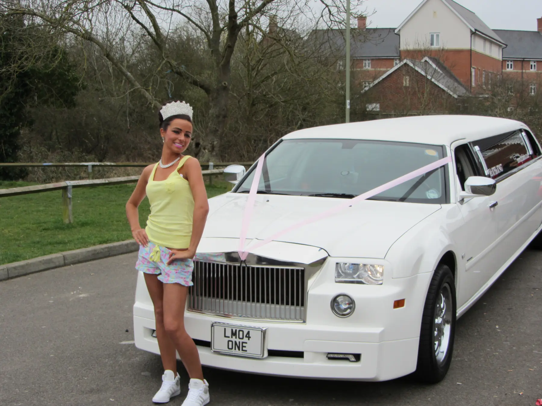 Young woman wearing crystal tiara posing in front of white Rolls-Royce style stretch limousine